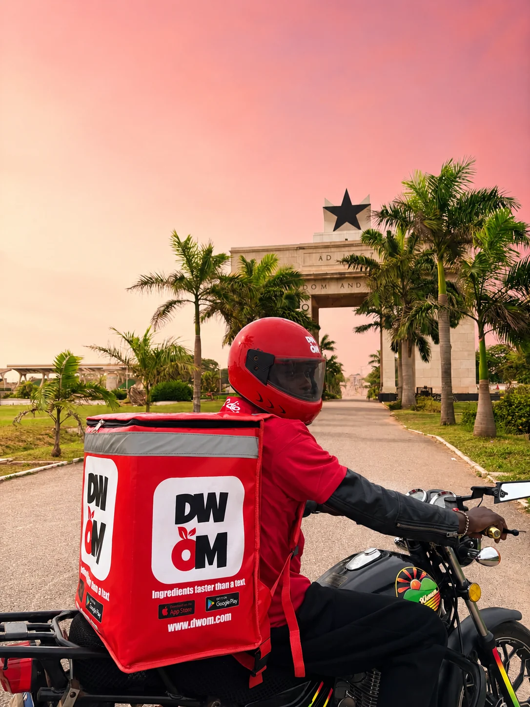 DWOM rider with a branded delivery box on a motorbike at Independence Arch, Accra, at sunset